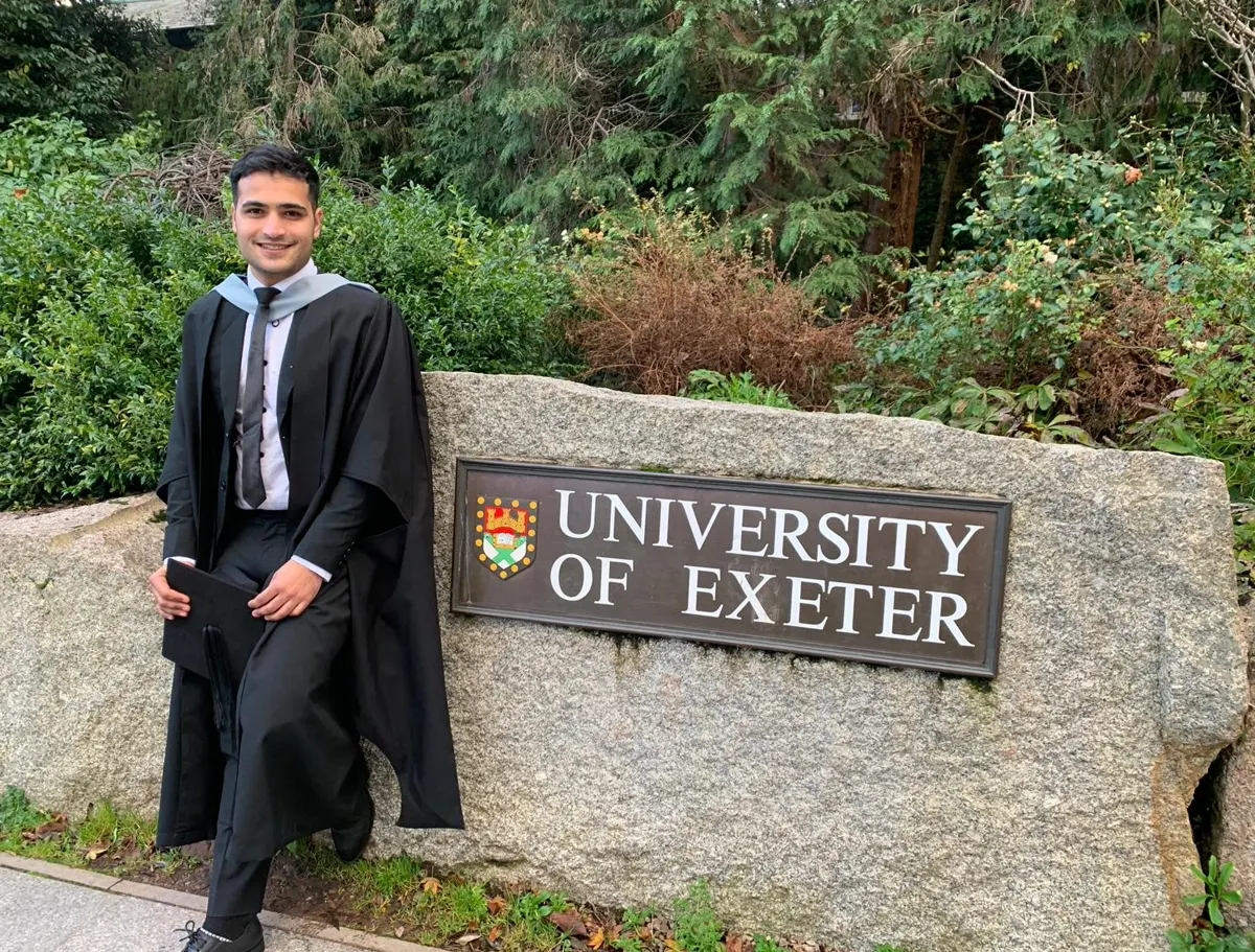 Subash in graduation gown beside the University of Exeter plaque