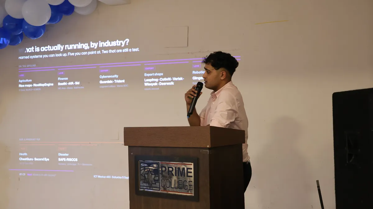 Subash speaking at a Prime College podium, holding a microphone in front of a projected slide